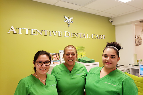 The image shows three individuals standing in front of a sign with the words  Attentive Dental Care  displayed on a green background, indicating they are at an office related to dental care services.