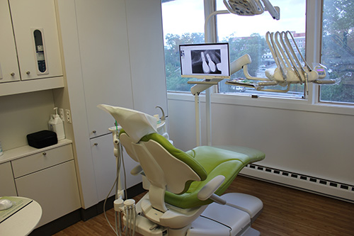 The image shows an interior view of a dental office with modern equipment, including a dental chair, digital display, and a window allowing natural light into the room.