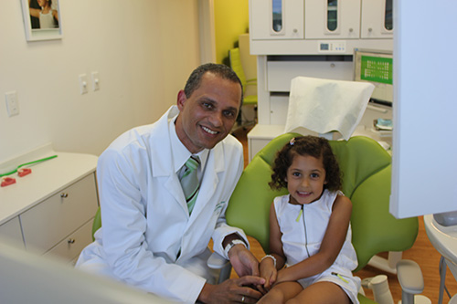 A dentist with a young child in his dental office, both smiling at the camera.