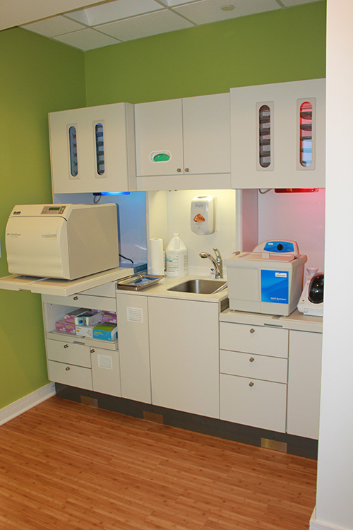 The image shows an interior view of a medical office with a clean and organized appearance, featuring cabinets, a sink, a countertop, and various pieces of equipment such as a printer and a microscope.