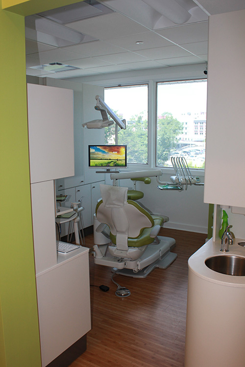 The image shows an interior view of a dental office equipped with modern dental chairs and equipment, featuring a green color scheme on the walls and a white floor, with a window revealing an outdoor scene.