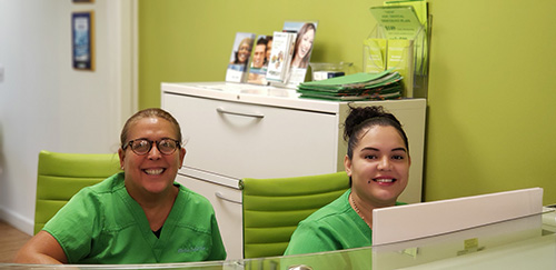 Two individuals seated at a reception desk with a green wall backdrop.