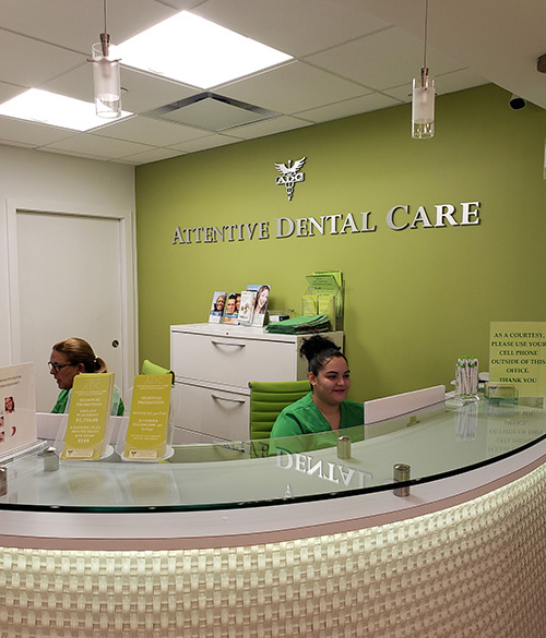 The image shows an interior view of a dental care office reception area with a sign indicating  Attentive Dental Care  on a green wall, a woman seated at the counter, and a desk with a computer monitor.