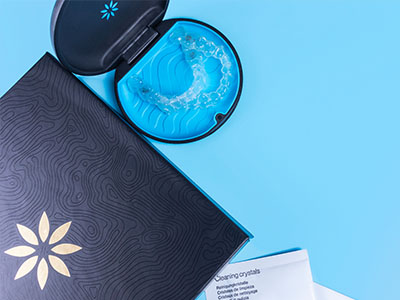 The image shows a blue toothbrush placed on top of a black book with a white cover featuring a floral pattern, next to a small, open box containing a dental cleaning product.