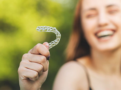 An adult woman smiling at the camera holds up a clear plastic retainer with her left hand.