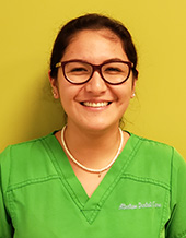 The image shows a person standing against a yellow background, wearing glasses, a necklace, and a name tag with text that is too small to read clearly. They are dressed in scrubs, which suggests they may be a medical professional such as a nurse or doctor.