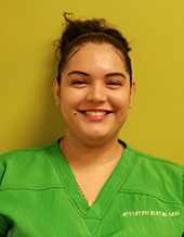 The image shows a smiling woman wearing scrubs, standing against a yellow background with a green wall on her left side.