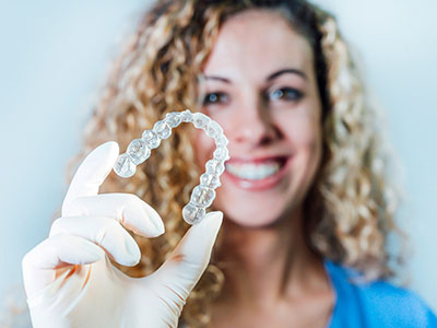 A woman wearing a blue shirt, smiling, holding up an oversized transparent dental implant with her left hand, while wearing white gloves and standing against a plain background.