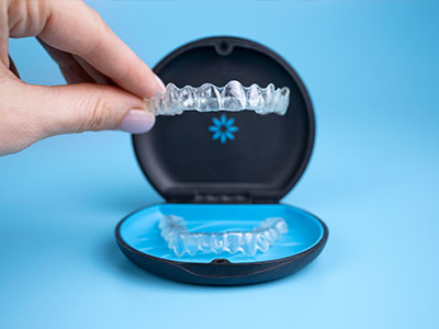 The image shows a person s hand holding a clear plastic tray with a set of transparent aligners inside, which are typically used for orthodontic treatment like Invisalign. The aligners are displayed in a blue and black case, suggesting they are being presented as an option for dental care.