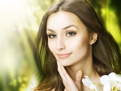 A woman with fair skin, looking directly at the camera, appears to be wearing makeup and has her hair styled down. She is set against a blurred background that suggests a natural environment with greenery.