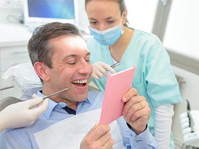 A man sitting in a dental chair holding up a pink card with a surprised expression while a dental professional looks on.