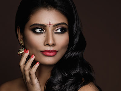 A woman with dark hair, wearing makeup, including red lipstick, and adorned with jewelry, poses for a portrait with her hand on her chin against a neutral background.