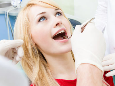 A woman seated in a dental chair receiving dental care, with a dentist performing a procedure on her teeth, visible dental instruments, and medical equipment.