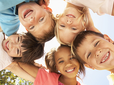 A group of children from diverse ethnic backgrounds posing happily with their arms around each other under a clear sky.