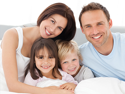 A family of four adults and three children posing together on a bed, with a smiling man and woman looking directly at the camera.