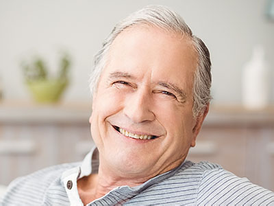 The image shows a smiling older man with gray hair, wearing a dark shirt, sitting comfortably with his arms crossed.