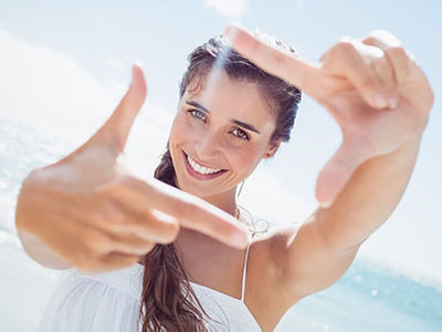 A woman with radiant skin is seen smiling at the camera while holding up a peace sign with her right hand. She has long, wavy hair and appears to be on a beach, given the bright sunlight and blue sky background.